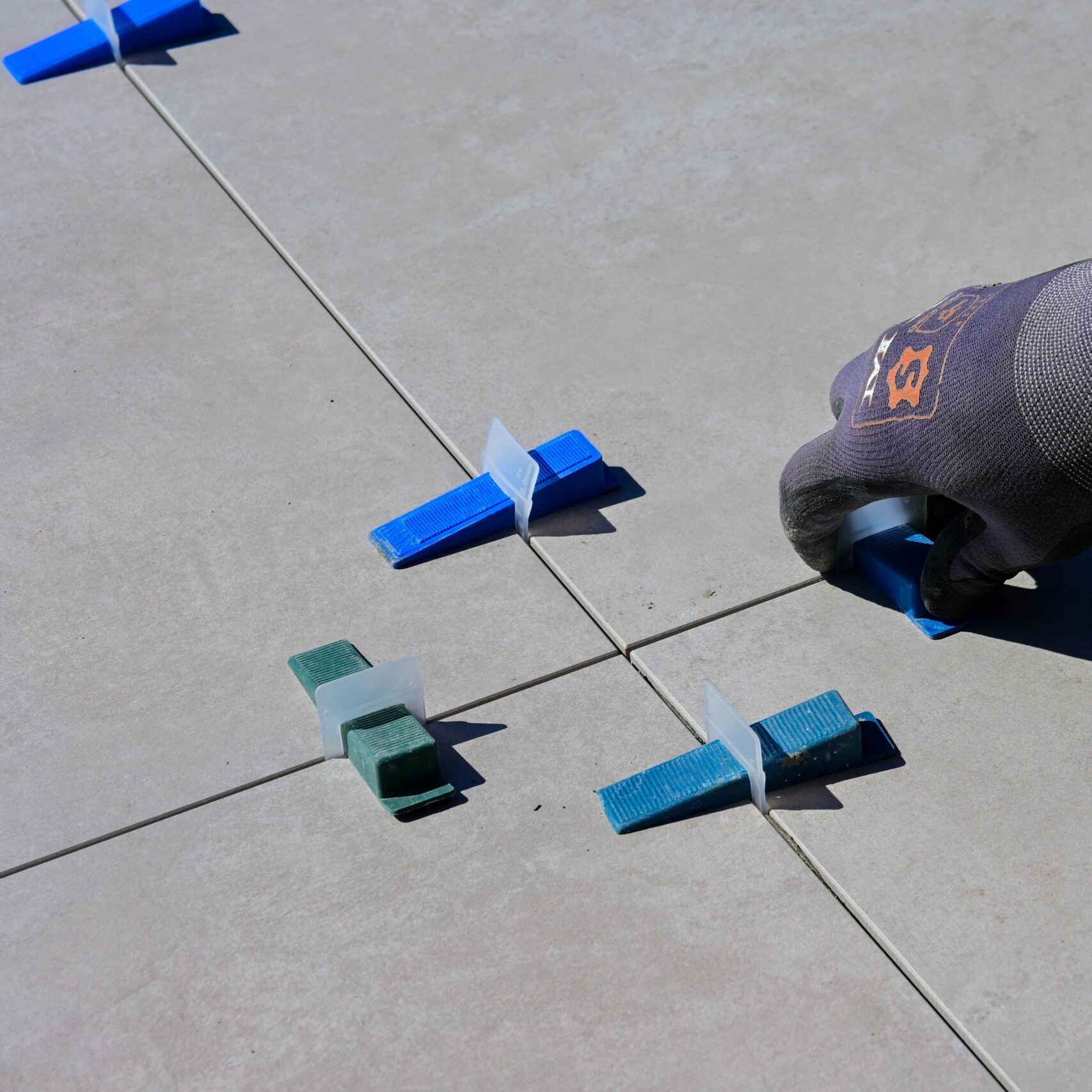 A close-up of a gloved hand installing tiles with spacers on a sidewalk.